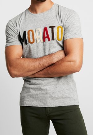 Uomo con le braccia incrociate che indossa una maglietta grigia con lettere multicolori e testurizzate che formano la parola "MORATO" e pantaloni verde scuro.