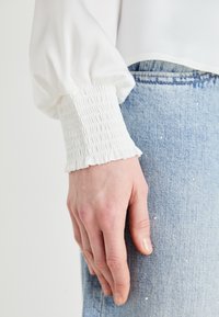 Blusa bianca con maniche a sbuffo e polsini arricciati, abbinata a denim azzurro chiaro con piccoli dettagli metallici. Materiale morbido e vestibilità comoda.