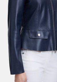 Marineblaue Lederjacke mit taillierter Passform, Reißverschluss vorne und praktischen Druckknopf-Taschen, kombiniert mit einer weißen Denim-Hose. Glatte Textur.