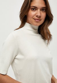 Suéter blanco de manga corta con cuello alto y textura suave. Presenta detalles de costura sutiles a lo largo de las mangas.