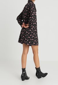 Vestido negro con flores y mangas largas, que presenta un corsé ajustado y un dobladillo acampanado. Combinado con botas de tobillo de charol negro y un detalle de cremallera.