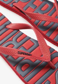 Tongs rouges et bleues avec des semelles en caoutchouc texturé. Présentent le logo "SUPERDRY" sur les sangles. Le design inclut des sangles larges et flexibles ainsi qu'un semelle à motifs.