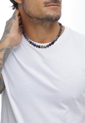 Collier en perles bleu et marron avec un pendentif en forme de crâne au centre, porté sur une chemise blanche unie. Perles lisses et rondes de textures variées.