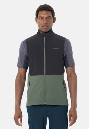 Uomo che indossa un gilet con zip nero e verde sopra una maglietta grigia a maniche corte, in piedi con le braccia rilassate contro uno sfondo bianco.