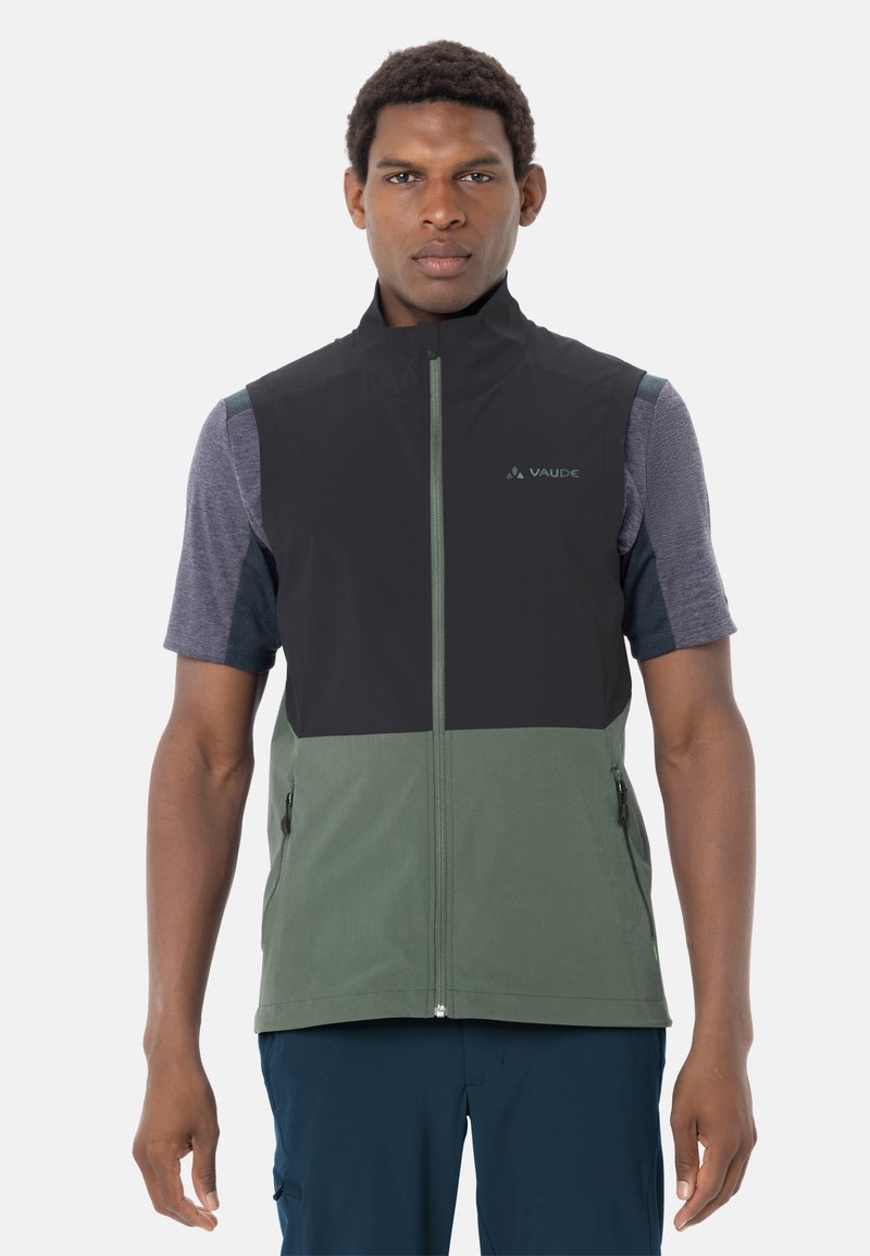 Mann trägt eine schwarze und grüne Weste mit Reißverschluss über einem grauen kurzärmligen Shirt und steht mit entspannten Armen vor weißem Hintergrund.