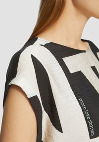 Zwart-witte grafische T-shirt met korte mouwen en een gekreukeld textuur. Kenmerkt zich door gedurfde geometrische patronen en subtiele turkooisaccenten. Tekst leest "meer liefdesverhalen."