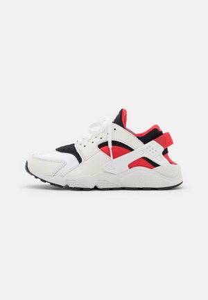 Chaussure de sport blanche avec des accents noirs et rouges, une semelle en caoutchouc texturée et un système de laçage. Des matériaux en mailles et synthétiques sont utilisés.