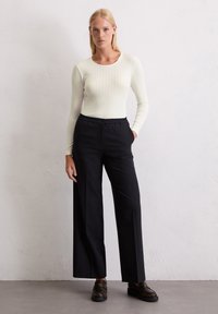 Femme se tenant contre un mur blanc, portant un haut long à manches longues blanc côtelé, un pantalon noir à jambes larges et des mocassins marron foncé.