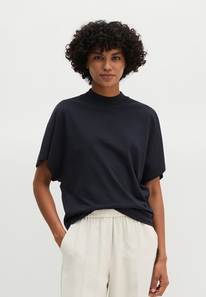 Frau mit lockigem Haar, die ein schwarzes kurzärmeliges Oberteil und hellbeige Hose trägt, mit Händen in den Taschen vor einem einfarbigen Hintergrund.