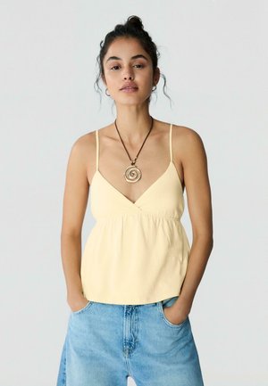 Ženska, oblečena v svetlo rumeno top brez rokavov in modre kavbojke, z rokami v žepih, nosi okroglo spiralno ogrlico.