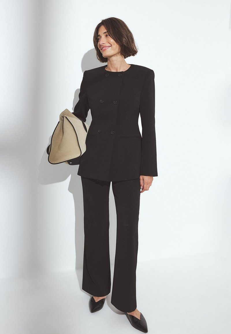 Black tailored double-breasted suit with structured shoulders, featuring long sleeves and flared trousers; complemented by a beige bag.