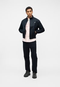 Άνδρας με μαύρο μπουφάν πάνω από ανοιχτό ροζ πουκάμισο, μαύρο παντελόνι και μαύρα αθλητικά παπούτσια, στέκεται με τα χέρια στις τσέπες του μπουφάν μπροστά σε λευκό φόντο.