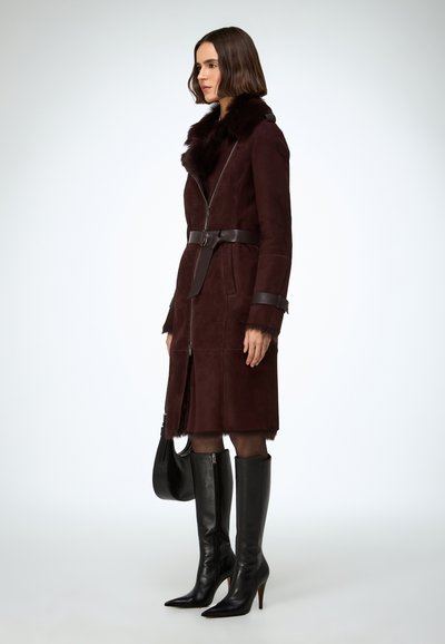 Kabát z bordovej semišovej kože s kožušinovým golierom a koženým opaskom, so zipsom na zapínanie a dlhými rukávmi. Modelka má na sebe čierne čižmy s vysokými podpätkami po kolená.