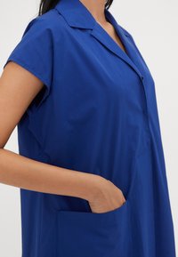 Donna che indossa un vestito camicia blu a maniche corte e larghe con colletto, mentre ha una mano poggiata in una tasca anteriore.