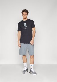 T-shirt Nike cinza escuro com logotipo, combinado com calções cinza claro. O modelo usa meias brancas e ténis com padrão preto e branco.