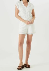 Biały kombinezon z ażurowej tkaniny z krótkimi rękawami, z przodu na guziki i z elastycznym paskiem w talii. Zestawiony z brązowymi sandałami na platformie.