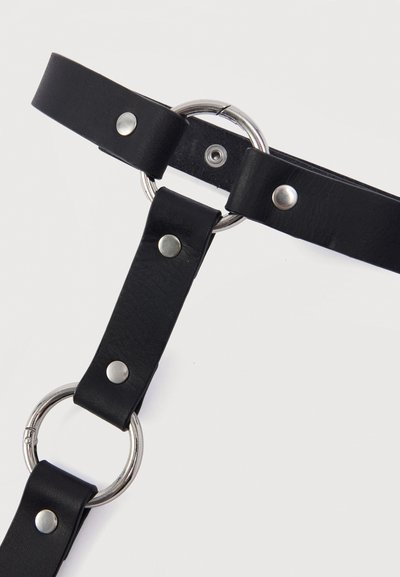 Μαύρα δερμάτινα λουριά ενωμένα με μεταλλικούς κρίκους και ασημένια καρφιά πάνω σε απλό λευκό φόντο.