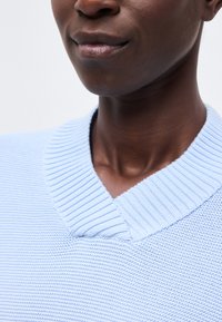 Gros plan sur la partie inférieure du visage et le cou d'une personne portant un pull en maille côtelée bleu clair avec un col en V.