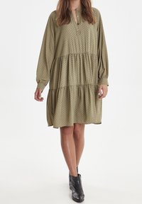 Olivově zelené šaty s dlouhými rukávy, knoflíkovou légou, volánkovým designem a černými puntíky. Nosené s černými kotníkovými botami.