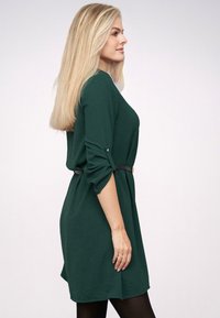 Grünes Jersey-Kleid mit Dreiviertelärmeln, einem schwarzen Gürtel auf Taillenhöhe und lässigem Schnitt. Glatte Textur und knielanges Design.