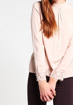 Woman wearing a pale pink long-sleeve blouse with lace cuffs and black pants, standing with hands clasped in front.