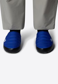 Pantofole imbottite blu con una suola nera testurizzata. La parte superiore presenta cuciture orizzontali e un logo sul lato.