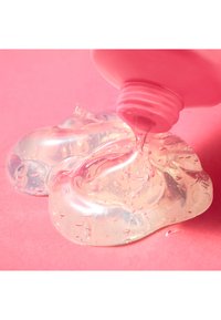 Un gel trasparente sta colando da una bottiglia con tappo rosa, poggiata su uno sfondo rosa. Il gel ha una consistenza liscia con piccole bolle visibili.