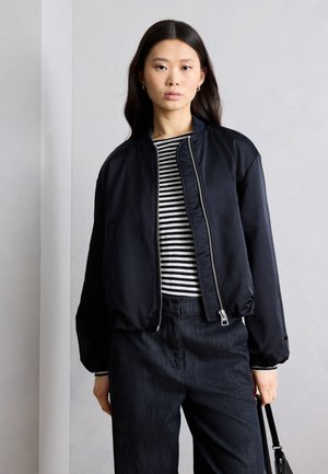 Ženska, oblečena v črno bomber jakno preko črne in bele črtaste majice ter temnih hlač, drži črno torbo ob sivi steni.