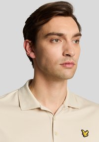 Beige polo-skjorte med teksturert design, med en tre-knappers placket og en gul fuglelogo på venstre brystområde.