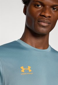 T-shirt de sport bleu clair avec un col rond et des manches courtes. Présente un logo Under Armour orange sur la poitrine. Matériau synthétique lisse.