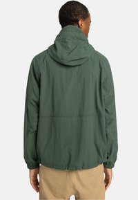 Grüne leichte Jacke mit Kapuze, elastischen Bündchen und lockerer Passform. Aus glattem Stoff mit einer leicht zerknitterten Textur gefertigt.
