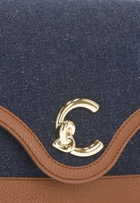 Borsa in denim con accenti in pelle color cuoio, caratterizzata da un design curvo e da una chiusura con logo dorato. Texture liscia con una leggera lucentezza.