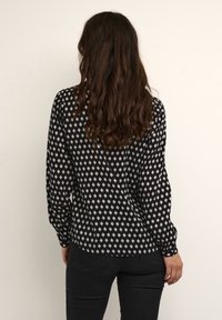 Schwarze Bluse mit langen Ärmeln und weißem Rautenmuster, mit gerundetem Saum und Knopfmanschetten, aus einem leichten Stoff gefertigt.