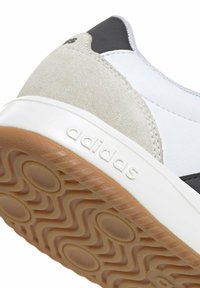 Primer plano del talón de una zapatilla Adidas que muestra gamuza gris, entresuela blanca con el logo de Adidas y suela de goma color marrón con patrón de dibujo circular y ondulado.