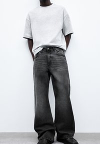 Grå oversize t-shirt og sorte jeans med vide ben. T-shirten har korte ærmer; jeansene har subtil falming og knapgylp. Fodtøjet er sorte sko.