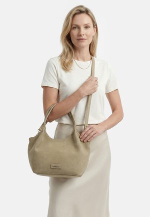 Vrouw draagt een wit T-shirt en een beige rok en houdt een beige handtas met twee hengsels vast tegen een effen witte achtergrond.