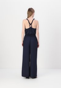 Marineblauer Jumpsuit mit einem taillierten Bund und weiter Beinoptik. Verfügt über einen verdrehten Rückenriemen mit einem runden Ausschnitt. Glatte Stofftextur.