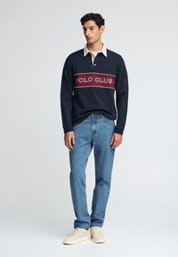 Granatowa koszula polo z długim rękawem, beżowym kołnierzykiem i bordowym paskiem z logo "POLO CLUB". Połączona z niebieskimi dżinsami i beżowymi sneakersami.