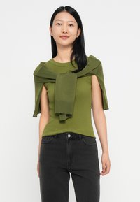 Jonge vrouw met steil zwart haar, gekleed in een olijfgroene mouwloze top en een bijpassende trui over haar schouders, gecombineerd met zwarte jeans.