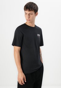 Ung mand med kort hår, iført en sort t-shirt med teksten "EMPIRE", står med den ene hånd i lommen foran en hvid baggrund.