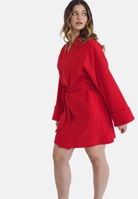 Czerwona, do kolan szata z teksturowanej tkaniny. Posiada zapięcie typu wrap, szerokie rękawy oraz wiązany pas w talii dla regulowanego dopasowania.