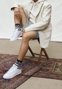 Valkoiset nahkakengät, joissa vihreät yksityiskohdat, yhdistetty harmaisiin shortseihin ja mustavalkoraidallisiin sukkiin. Istuva puisella tuolilla kuviollisen maton päällä.