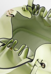 Vihreä synteettinen ämpäri laukku, jonka sisällä on rypytetyt reunat, musta reunus ja pyöreä kultainen lisäosa. Lempeä pinta, tilava muotoilu.