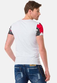 Weißes T-Shirt mit bunten gemusterten Ärmeln, schwarzen, roten und weißen Akzenten. Figurnahes Design mit glatter Textur. Getragen mit dunkelblauen Jeans.
