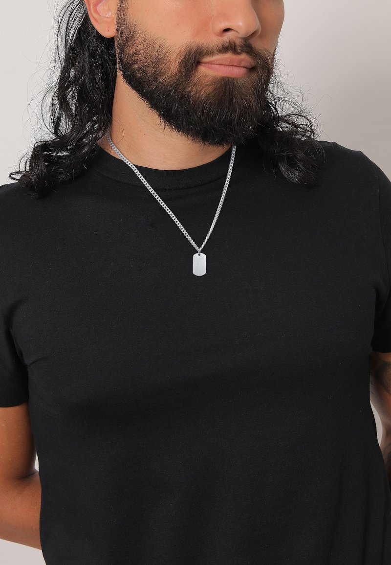 Collier en chaîne argentée avec un pendentif en plaque militaire porté sur un t-shirt noir uni. La chaîne présente un motif tressé et une texture lisse.