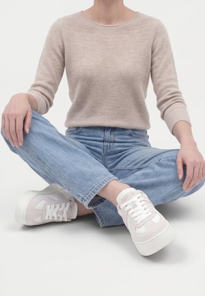 Person trägt beigen Pullover, hellblaue Jeans und weiße Sneakers und sitzt mit übergeschlagenen Beinen auf einer weißen Oberfläche.