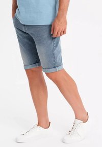 Shorts de mezclilla para hombre en azul claro, con dobladillos enrollados, corte recto y zapatillas blancas. Textura suave con un diseño clásico.