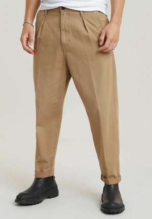 Beige chinos with a straight leg, featuring front pleats, belt loops, and rolled cuffs, paired with black ankle boots.