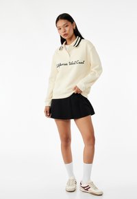 Kremowy sweatshirt z czarnymi akcentami, z napisem "California West Coast". W połączeniu z czarną spódnicą i białymi skarpetkami do kostek.