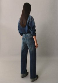 Chaqueta de mezclilla y jeans azules de tiro alto con un corte holgado. La tela está desgastada con un acabado texturizado, con puños enrollados y bolsillos traseros.